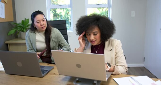 Diverse female coworkers reacting to laptop alert at desk, putting down phone and resolving issue