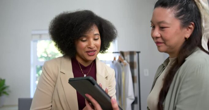 Diverse female duo holding tablet up, leaning in, pointing, reviewing outfits in studio with rack