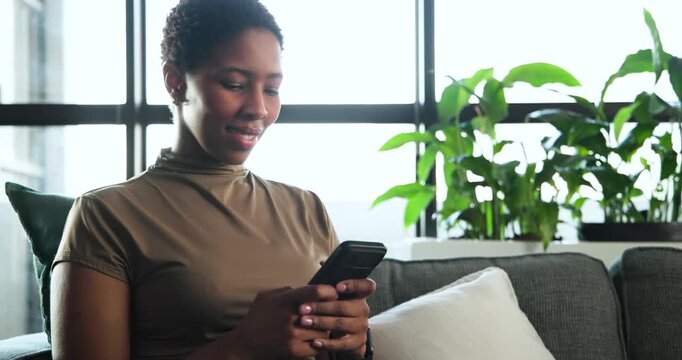 African woman scrolling smartphone and reading messages placing it on gray sofa in living room