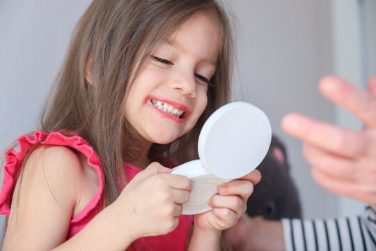 Smiling young girl with lipstick and compact mirror, enjoying playful makeup practice at home with soft light and blurred background, childhood beauty routine and self expression