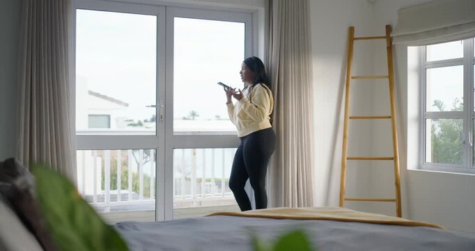 African American woman taking call, holding smartphone, speaking at sliding door in bedroom