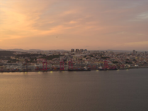 Aerial view of the Port of Lisbon with container cranes and cargo ships along the waterfront, set against the city skyline under a warm sunset sky in Lisbon, Lisbon, Portugal.