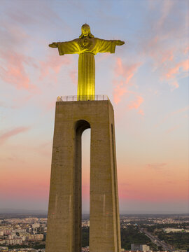 Aerial view of the illuminated Cristo Rei statue standing tall against a soft pink and orange sunset sky overlooking the urban landscape of Lisbon, Lisbon, Portugal.