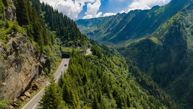 Aerial view of the Transfagarasan mountain road winding through lush green forests and rocky cliffs of the Carpathian Mountains in Curtea de Arges, Judetul Arges, Romania.