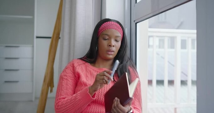 African American woman opening dark notebook and journaling on window seat, sipping mug, gazing out