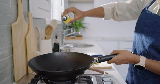 African American woman wearing blue apron at stove holding wok on lit burner pouring oil cooking
