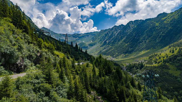 Aerial view of the Transfagarasan mountain road winding through lush green forests and rugged peaks under a blue sky with white clouds in Curtea de Arges, Judetul Arges, Romania.