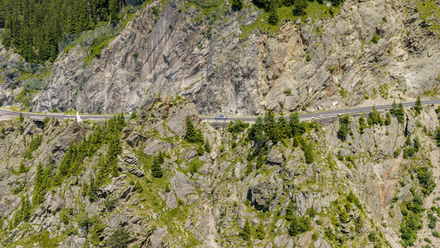 Aerial view of a car driving on the winding Transfagarasan mountain road carved into rocky cliffs, surrounded by steep slopes and pine trees, Curtea de Arges, Judetul Arges, Romania.
