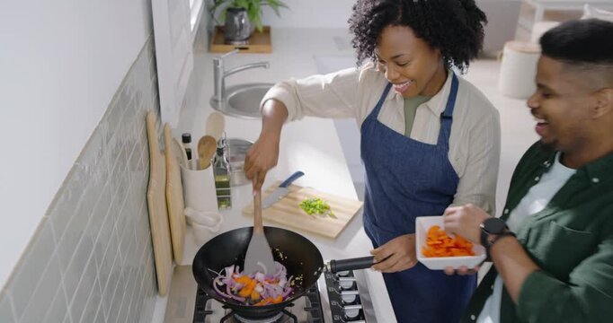 African American couple stirring wok, spatula at stove, man offering bowl adding carrot stir-fry