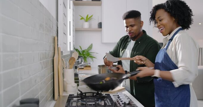 Couple cooking, woman stirring veg in wok in kitchen while man bringing bowl adding seasoning