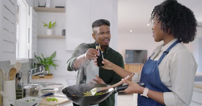 African American couple cooking in kitchen, stirring wok, man flinching from steam offering bottle