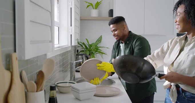 African American couple after meal cleaning at sink, man rinsing plates gloved, woman drying wok