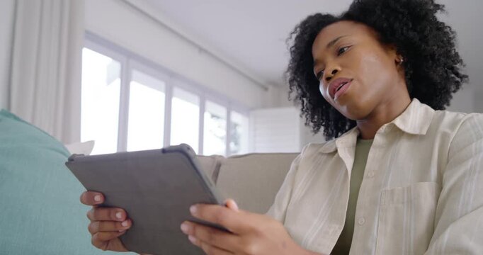 Adult African woman holding tablet on sofa by windows, studying then tapping screen for answers