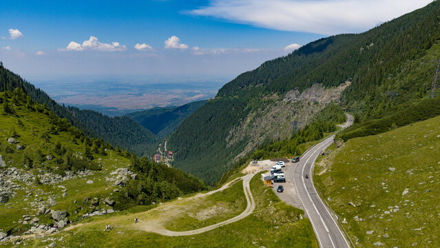 Aerial view of the Transfagarasan mountain road winding through the lush green Carpathian Mountains with parked cars and distant valley views in Curtea de Arges, Judetul Arges, Romania.