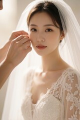 Fototapeta premium Young woman in white lace bridal gown gets ear piercing. Bride wears veil, looks at camera. Gentle hands adorn her with jewelry before wedding ceremony, soft light.