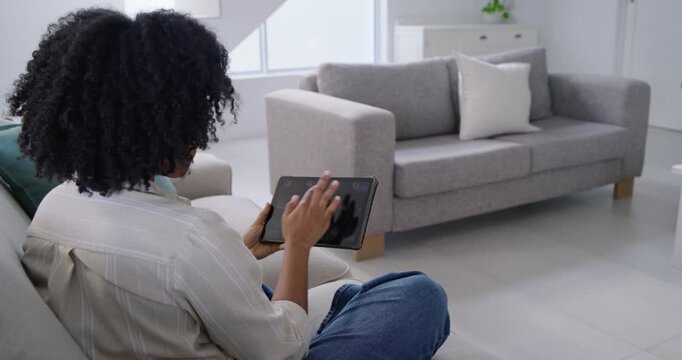 African American woman tilting tablet, tapping and swiping while choosing content on gray sofa