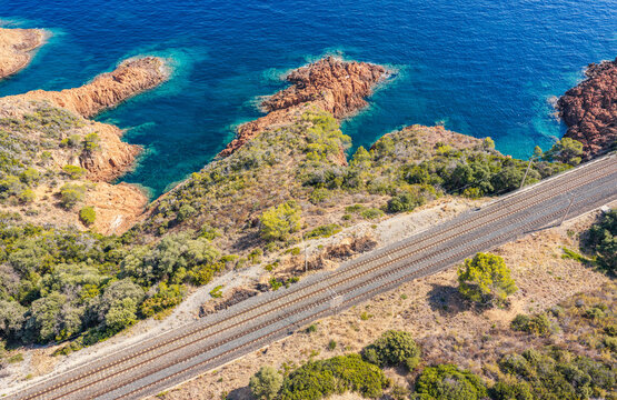Aerial view of an electrified railway track running along the rocky Mediterranean coastline with red rock formations and turquoise coves in Saint-Raphael, Provence-Alpes-Cote d'Azur, France.