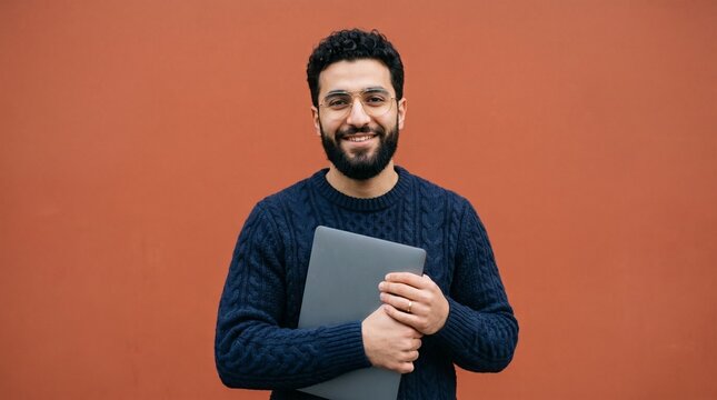 Cheerful young man with a thick dark beard wearing glasses holding a grey laptop while smiling against a terracotta wall