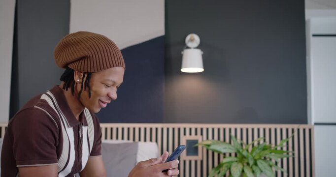 African American man sitting on couch holding blue phone, reading message and recording voice reply