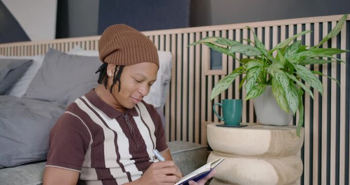 African man holding pen, writing blue notebook on couch, flipping it, sipping green mug, reflecting