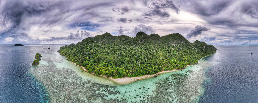 Aerial view of Raja Ampat Regency featuring a lush tropical island with dense forests, white sand beach, and vibrant coral reefs in turquoise waters under a cloudy sky in Southwest Papua, Indonesia.
