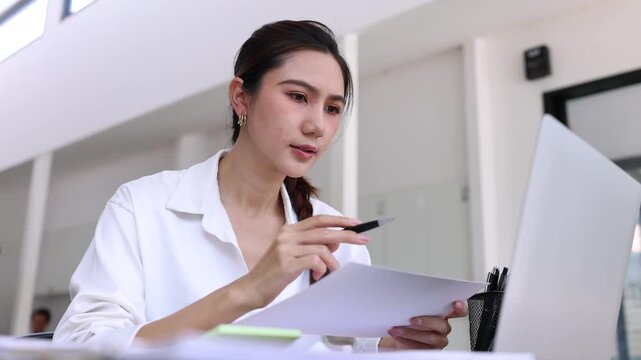 Asian businesswoman in a white shirt working in a modern office, sitting at her desk with a laptop, looking at and signing documents with a pen while reviewing business reports and data.