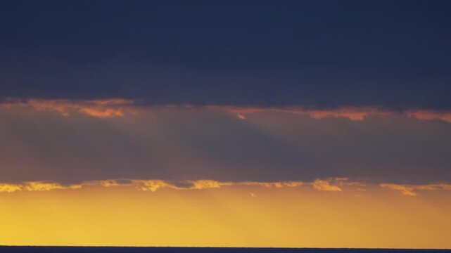 Golden sunbeams pierce dark heavy storm clouds above a calm sea surface at twilight. Static wide shot.