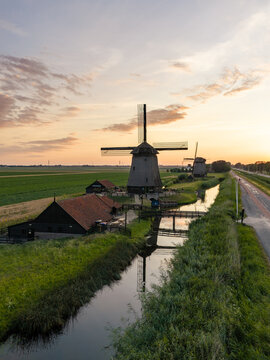 Aerial view of De Museummolen windmill surrounded by lush green fields, a calm canal, and traditional wooden buildings under a warm sunset sky in Schermerhorn, Noord-Holland, Netherlands.