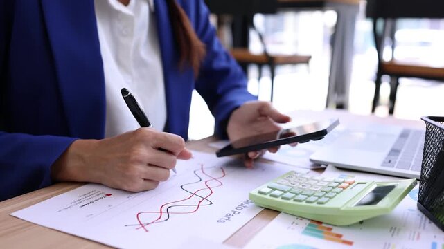 Asian businesswoman in a suit using a smartphone at her desk, surrounded by financial reports, charts, and a calculator in a modern office, Concept of corporate work, mobile banking and communication.