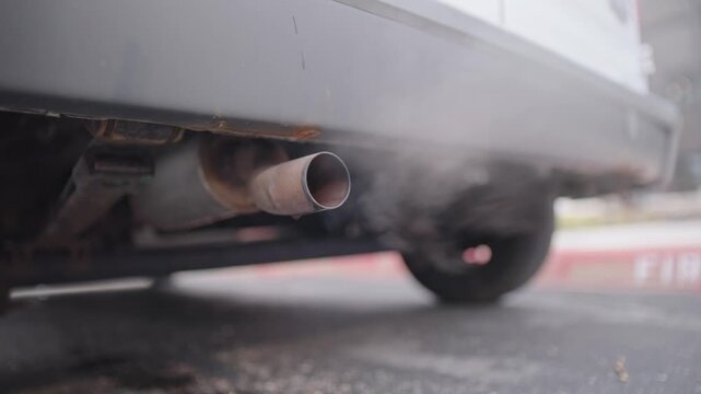 Water vapor, smoke, and steam shoot and exit out of old rusty van car truck exhaust pipe system connected to older muffler pipes beneath back of white aged vehicle during startup of maintenance 4k