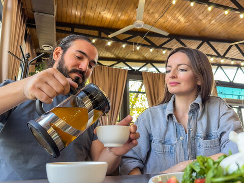 Man pouring hot tea from glass teapot into cup beside woman seated at restaurant table, relaxed indoor dining scene with warm wooden ceiling, soft daylight, casual conversation, beverage service