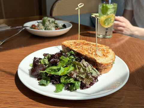 Grilled bread and mixed greens on white plate beside citrus mint drink, with second salad plate and soft diner background in sunlit restaurant table setting, with food, restaurant, salad, bread detail