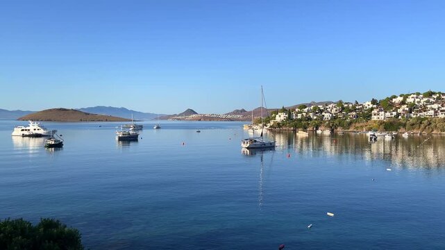 Scenic view of the calm, blue bay in Bitez, Turkey, with luxury boats anchored on the still water near a hillside resort town under a clear morning sky.