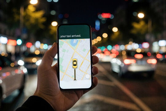 Hand holds smartphone with taxi app showing map, car location. Mobile phone screen displays arrival notification on dark city street. Blurred urban night background with bokeh lights from traffic,