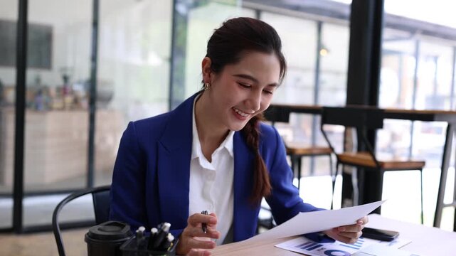 A smiling young Asian businesswoman wearing a blue blazer working in a modern office, happily reading and reviewing a financial report document at her desk with a laptop.