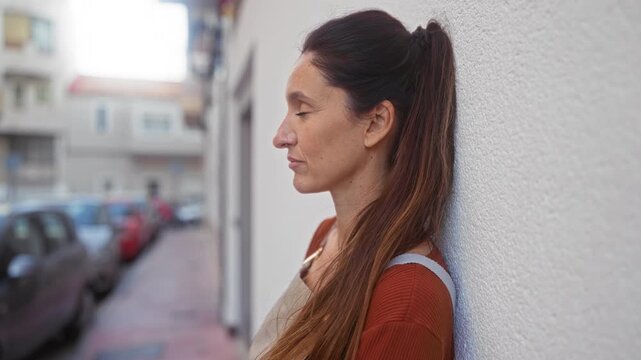 Woman leaning against a white wall on street, apron strap visible and face in profile, eyes closed and shoulder pressed to wall; quiet contemplation.