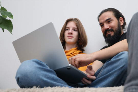Young person and bearded man sharing laptop and tablet indoors, seated on floor beside houseplant with relaxed focus, simple home setting, low angle composition, casual learning moment