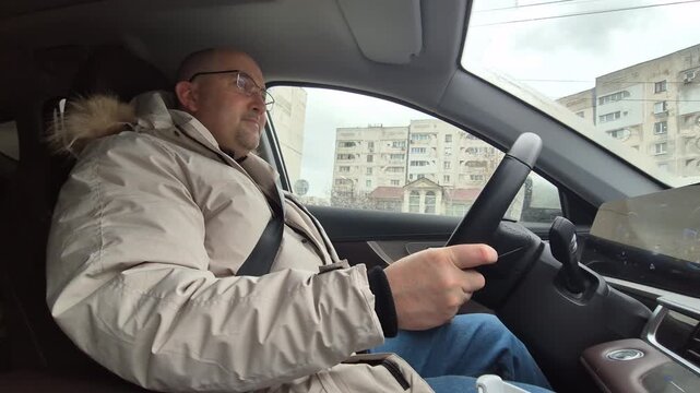 Adult man driving car through city streets while wearing winter jacket. Static side profile shot.