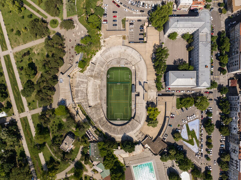 Aerial view of Tasmajdan Stadium featuring a green soccer pitch surrounded by concrete seating, adjacent to park areas, parking lots, and urban buildings in Beograd, Grad Beograd, Serbia.