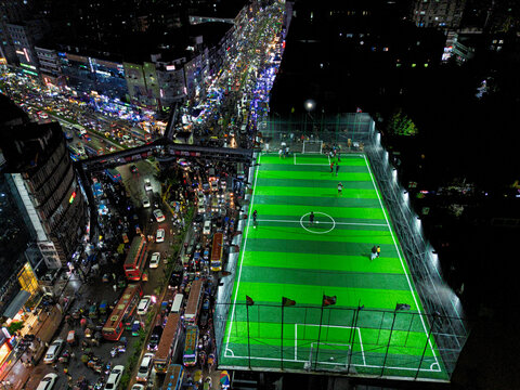 Aerial view of a vibrant rooftop football pitch illuminated at night, situated beside a bustling city street filled with traffic in Dhaka, Dhaka Division, Bangladesh.