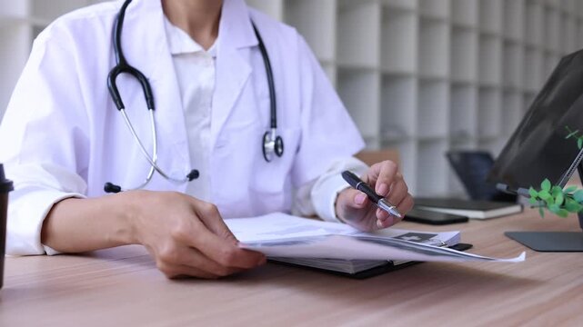 Asian female doctor using digital tablet for online video call consultation with patient and reading medical report in hospital office.
