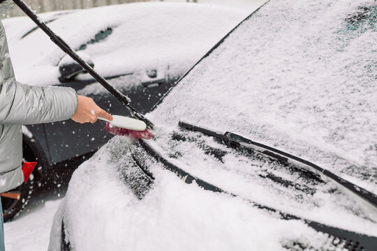 Close-up of a person clearing fresh white snow off an automobile front window and wipers after blizzard. Winter vehicle maintenance, automotive care, and freezing weather concept.