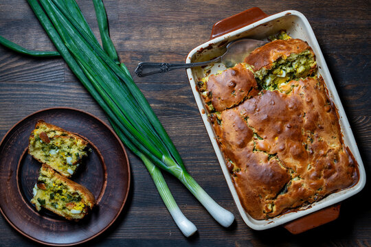 Homemade green onion and egg pie in baking dish on wooden table, closeup. Savory scallion and egg casserole pie served in ceramic baking dish with sliced pieces on plate and fresh green onions nearby