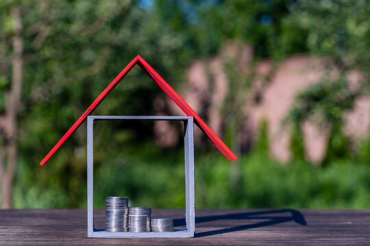 House model with red roof and coin stacks inside. Simple wooden house frame with bright red roof and three stacks of silver coins inside stands on wooden surface against green garden background