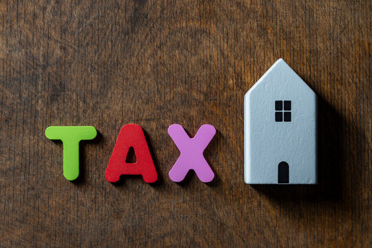 Property tax concept with word tax and wooden house model, closeup. The word TAX in colorful wooden letters placed next to small white toy house model on rustic dark wooden background