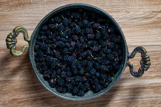 Top view of fresh black mulberries in vintage metal bowl, closeup. Flat lay shot of rustic metal bowl with ornate handles filled with ripe black mulberries on natural wooden table surface