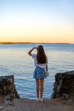 Woman Looking Across Calm Water at Sunset. Woman standing on a rocky shore, looking across calm blue water toward the horizon at sunset.