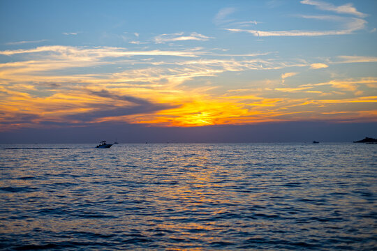 Golden Sunset Over Calm Sea with Boats. Orange sunset over calm sea with layered clouds, gentle waves, reflections and small boats silhouetted near the horizon.