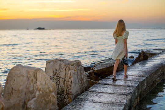 Young Girl Walking by the Sea at Sunset. Young girl in a summer dress walking along a stone pier beside the sea at sunset.