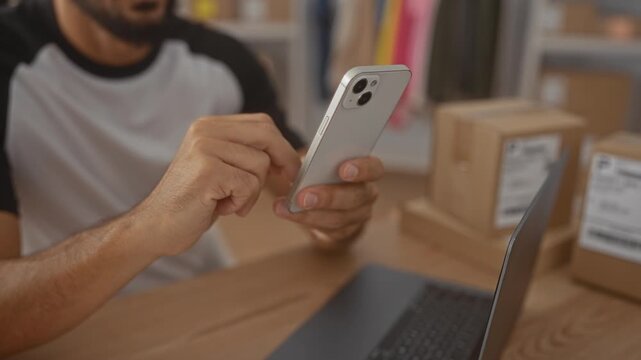 Man tapping smartphone with fingers at packing desk in building, checking orders beside laptop and shipping boxes; small business efficiency focused.